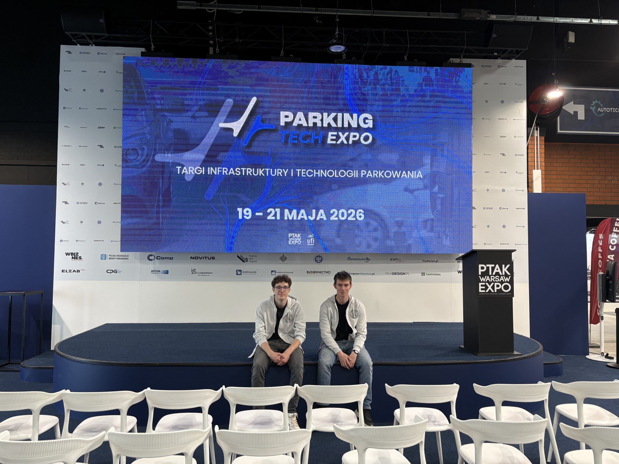 Zdjęcie dwóch mężczyzn, Jana Polaszka (Lidera zespołu) oraz Adama Kolosę, uczestników wydarzenia ParkingTechEXPO. Nad mężczyznami wisi duży cyfrowy ekran, na którym napisano: PARKING TECH EXPO TARGI INFRASTRUKTURY TECHNOLOGII PARKOWANIA 19-21 MAJA 2026.